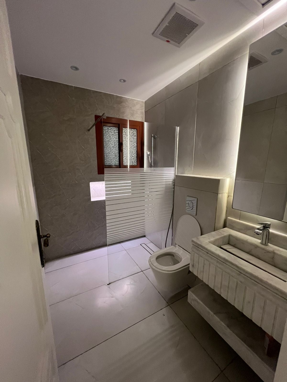 A modern bathroom features light-colored tiles and a large mirror reflecting natural light. A shower stall is separated by frosted glass. A sleek sink with a minimalist design sits atop a stone surface, beside a toilet, completing the space with a clean and functional layout.