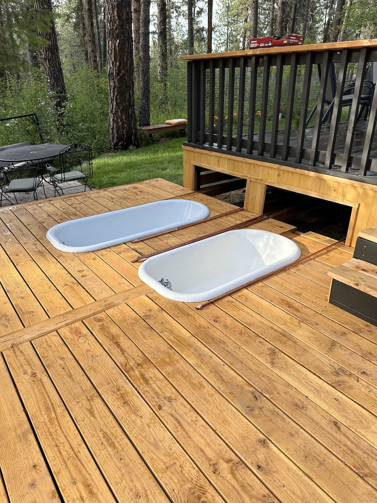 Two freestanding soaking tubs are positioned on a wooden deck, inviting relaxation in a natural setting. The deck is surrounded by lush greenery, and a shaded area of the deck features seating arrangements, enhancing the tranquil atmosphere.