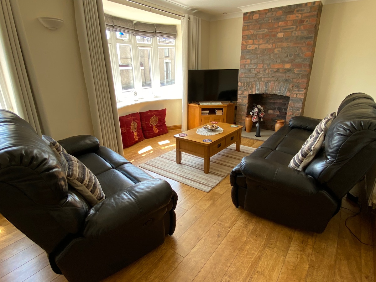 The living area features two reclining leather sofas facing a wooden coffee table, with a brick fireplace and modern television. Natural light enters through a large bay window adorned with curtains, while decorative cushions are placed on a low bench nearby.