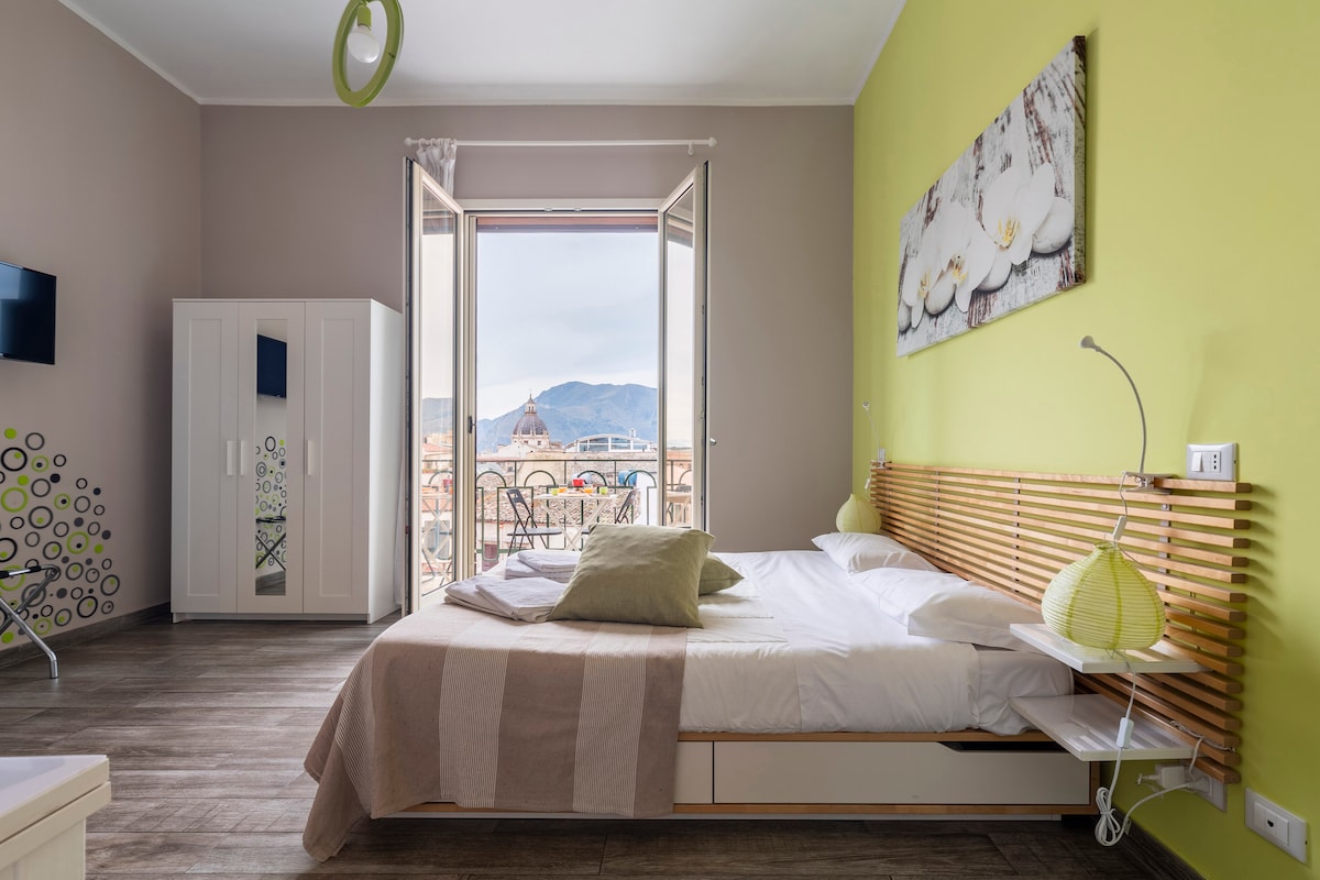 A light and airy bedroom features a spacious bed with a striped blanket, complemented by green accents on the walls. Large windows open to a balcony offering views of the surrounding landscape. A wardrobe and wall-mounted television add convenience to the space.