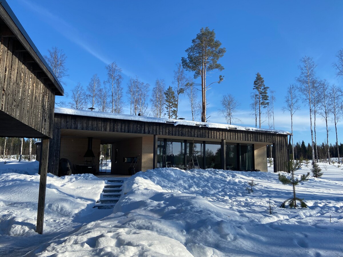 A modern cabin is depicted, showcasing a flat roof and wooden exterior. Snow covers the surrounding area, highlighting the natural landscape. Large glass windows provide a view into the interior space, while tall trees stand in the background against a clear blue sky.
