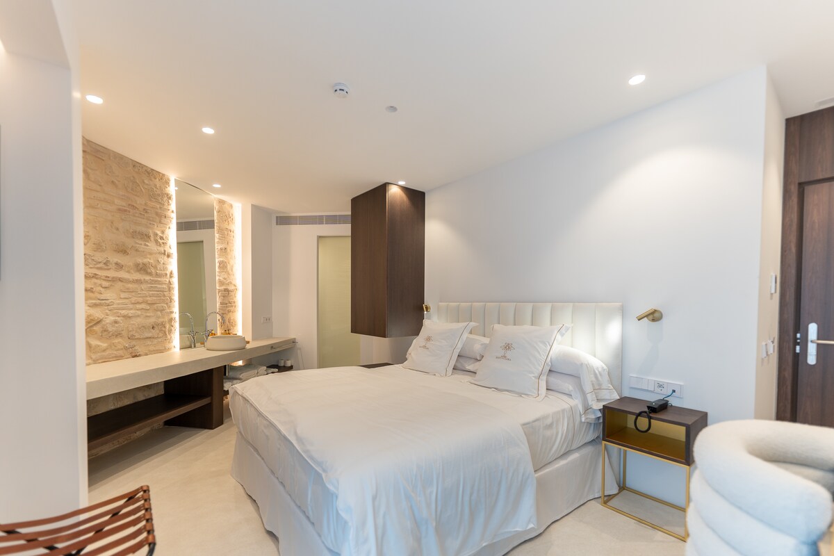 A modern bedroom is observed, featuring a spacious bed with light-colored bedding. A stylish bedside table and a softly illuminated wall accent add modern touches. A sleek vanity area is visible in the background, complemented by natural stone details, enhancing the room's contemporary design.