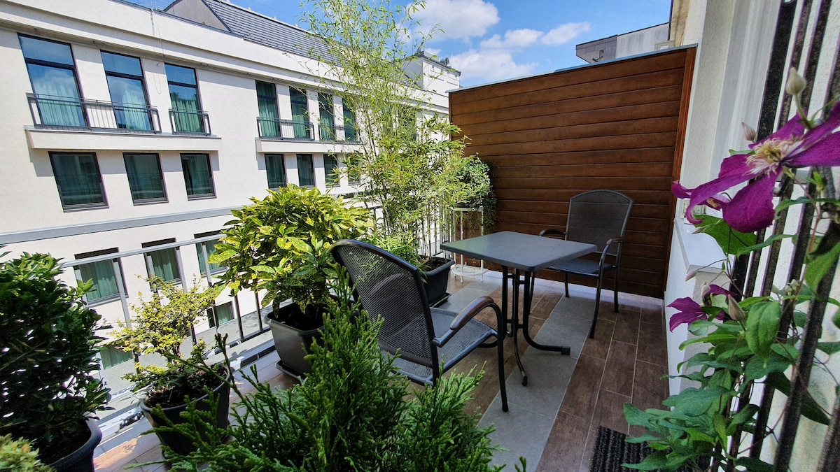 A private terrace is furnished with a table and chairs, surrounded by various potted plants. The space offers a view of buildings across the street, with greenery creating a tranquil environment. Sunlight brightens the area, enhancing the cozy outdoor seating.