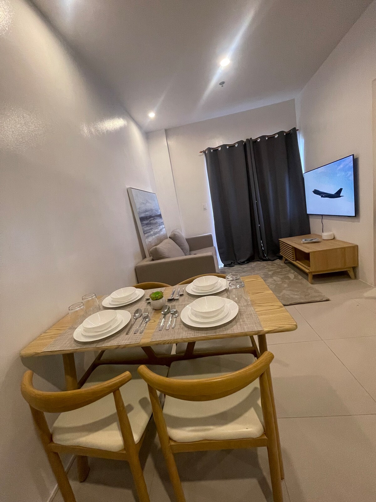 A dining area features a wooden table set for four, complemented by light-colored chairs. A cozy seating area with a neutral sofa is visible in the background, along with a television mounted on the wall. Dark curtains provide privacy while natural light brightens the space.