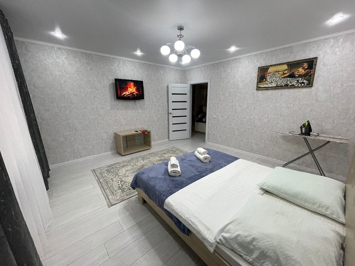 A spacious room features a light-colored bed with fresh linens and towels neatly arranged. A cozy ambiance is created by a wall-mounted television and decorative artwork. An ironing board stands adjacent, while soft lighting from a ceiling fixture enhances the overall calm atmosphere.