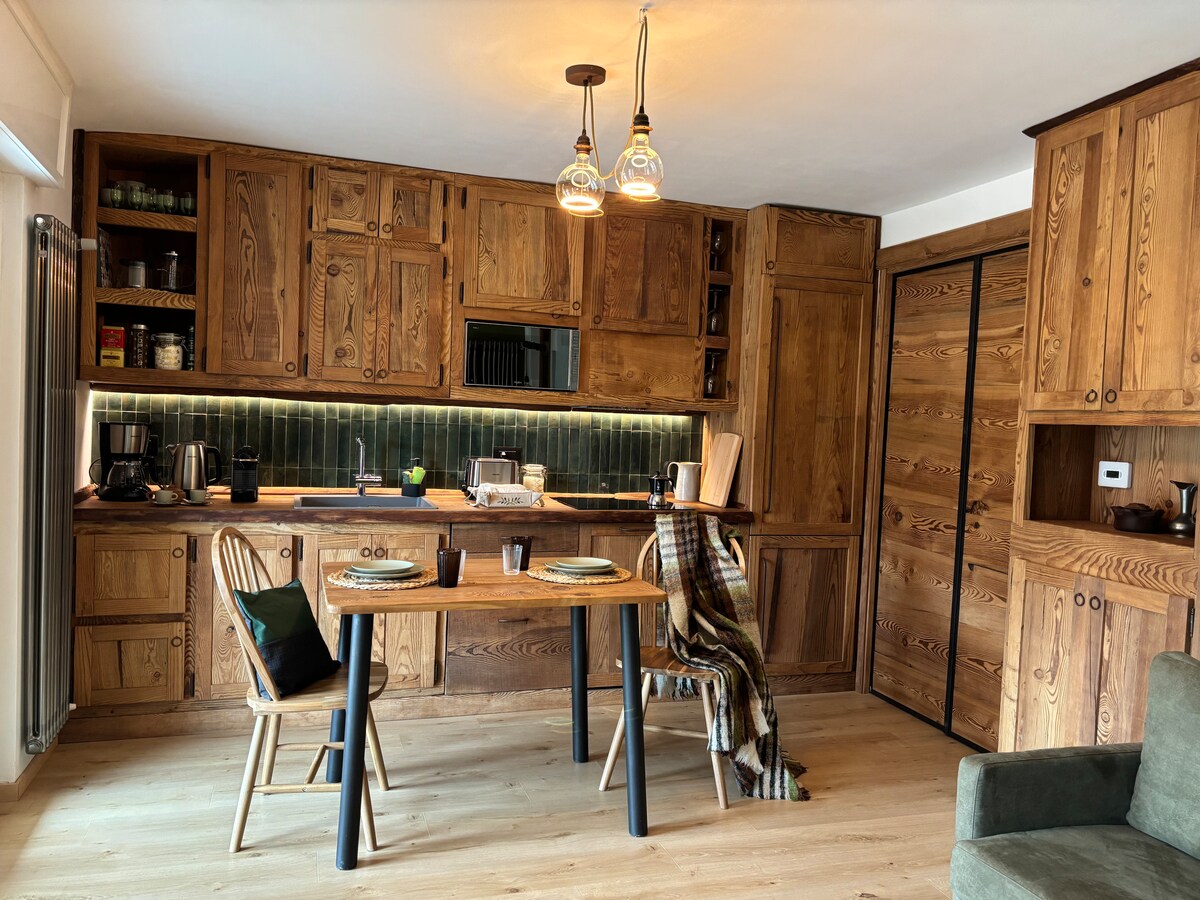 The kitchen features natural wood cabinetry and shelves, complemented by modern appliances. A dining table is set for two, surrounded by wooden chairs. Soft lighting is provided by pendant lights above, creating an inviting space for meal preparation and dining.