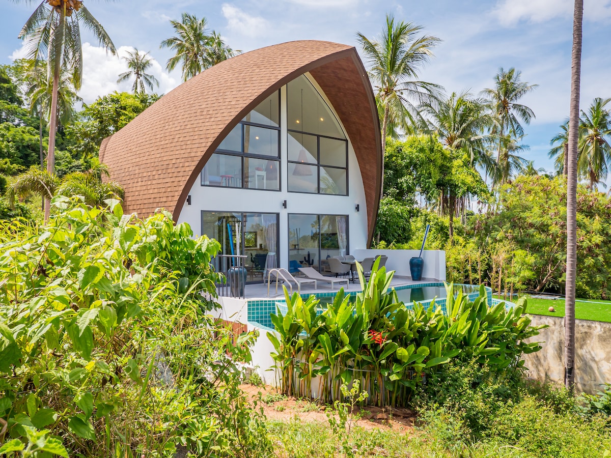 A unique villa is surrounded by lush greenery and tropical plants. Its curved roof and large glass windows offer a modern aesthetic. A pool is visible in the foreground, reflecting the sky and providing a serene outdoor area for relaxation.