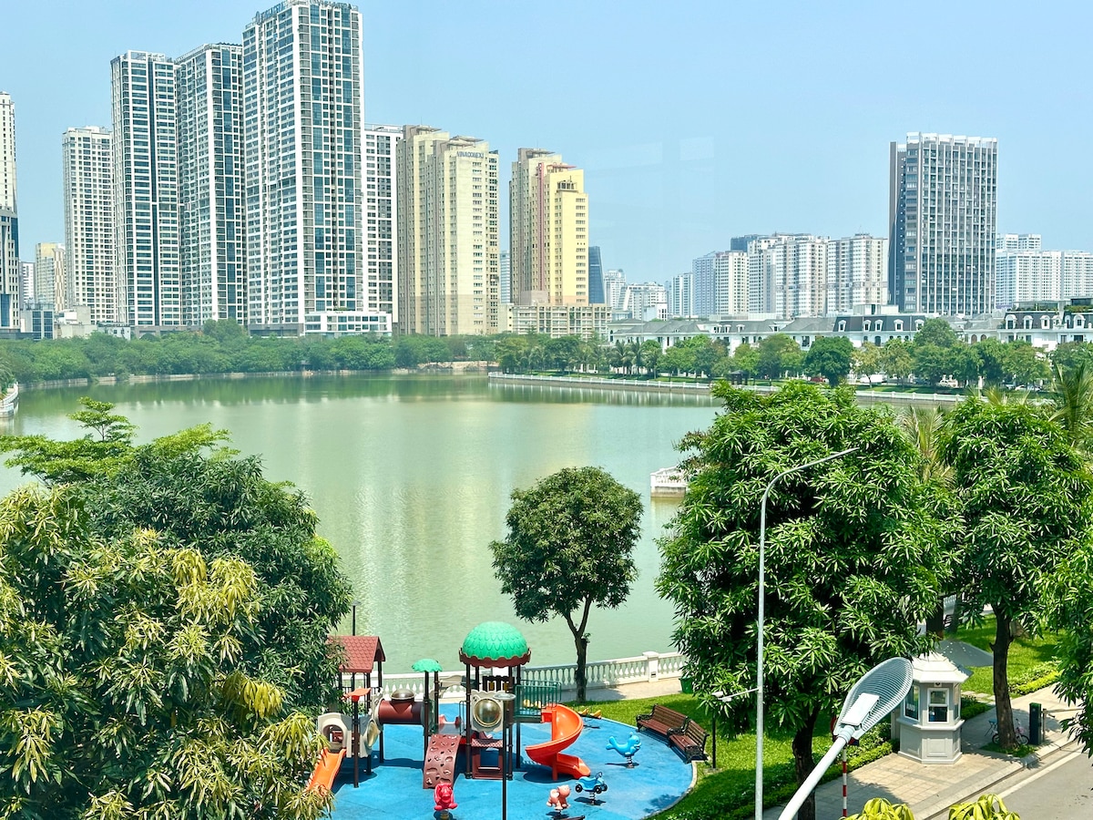A serene view of lush greenery and a serene lake is complemented by a children's playground featuring colorful slides and play structures. Modern high-rise buildings line the background, reflecting the vibrant urban environment of Vinhomes Green Bay.
