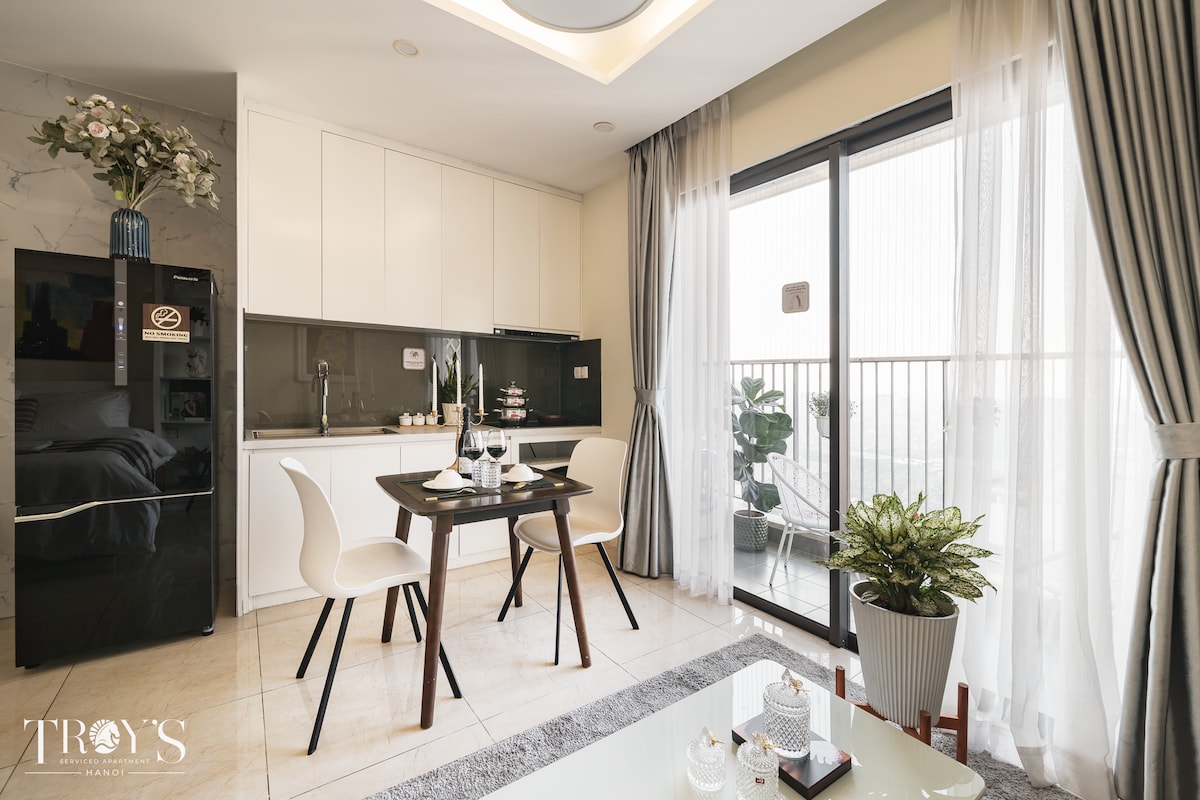 The image captures a modern kitchen and dining area, featuring a sleek black refrigerator, a small dining table with comfortable chairs, and a vibrant potted plant. Sunlight filters through large glass doors that open to a balcony, enhancing the inviting atmosphere of the space.
