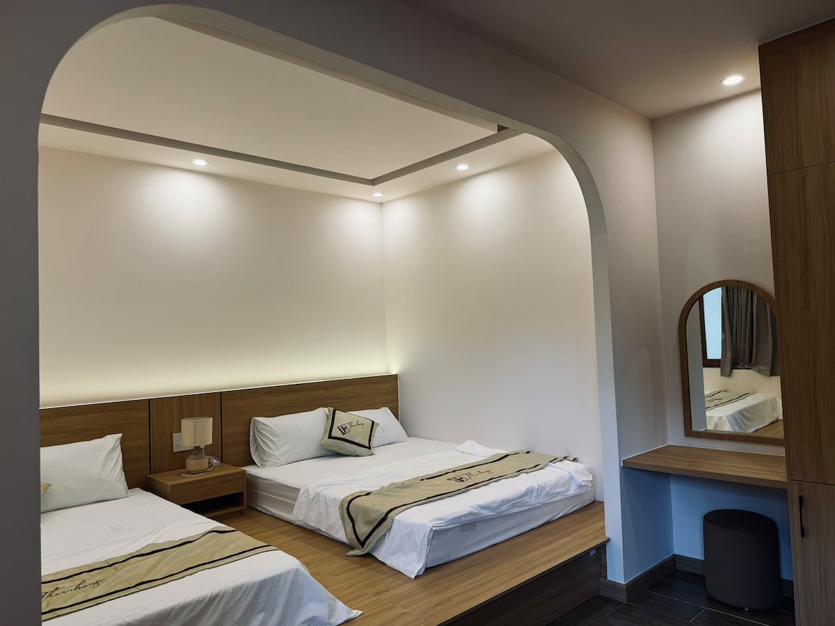 The room features two beds with understated linens placed against a softly lit backdrop. A wooden vanity with a round mirror is positioned to one side, while natural light streams in through a nearby window, accentuating the warm color palette.