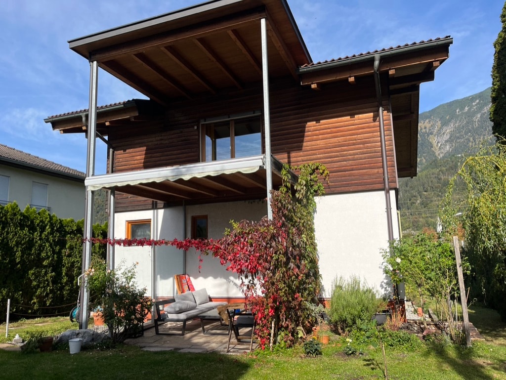 A two-story apartment is shown from the garden, featuring a covered terrace with seating. Climbing plants and greenery surround the structure, while scenic mountains rise in the background under a clear blue sky.