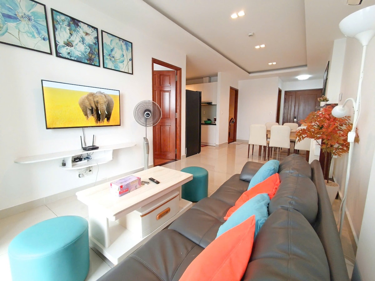A modern living area showcases a dark leather sofa adorned with colorful cushions. A large TV is mounted on the wall, displaying an image of elephants. Light-colored furniture and a decorative fan enhance the space, with a dining area visible in the background.