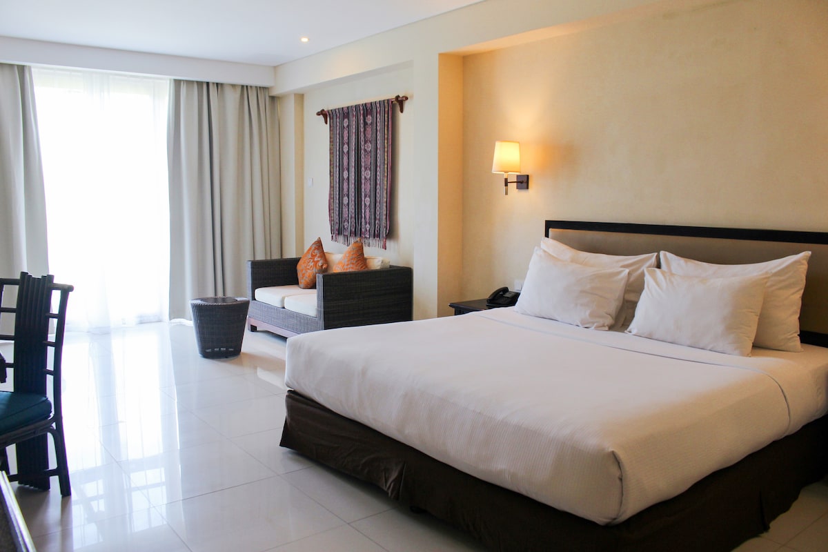 A spacious room is furnished with a king-sized bed, complemented by crisp white linens. A cozy seating area features a sofa with decorative cushions. Natural light enters through large windows, and a small table is arranged nearby.