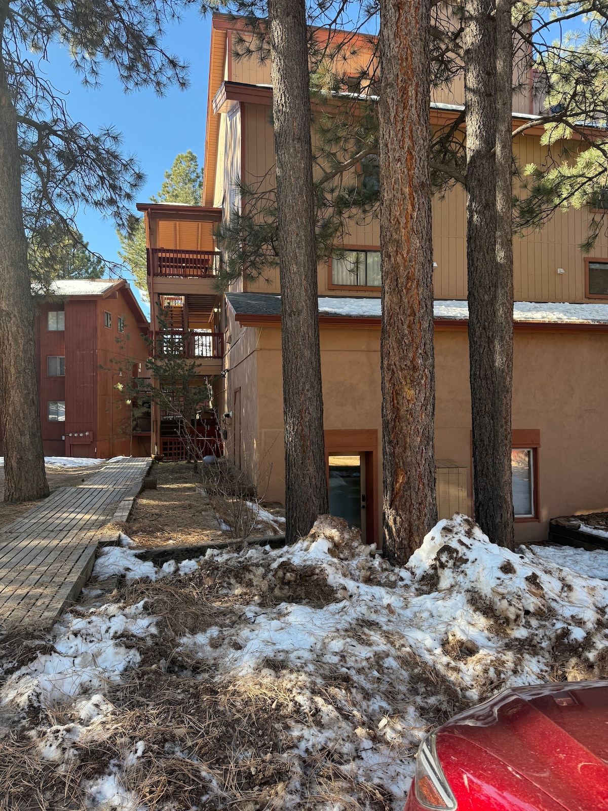 The exterior of a two-story condo is visible, surrounded by tall pine trees. Snow is present on the ground, indicating winter conditions. A walkway leads to the entrance, while another building can be seen in the background, enhancing the complex's character.