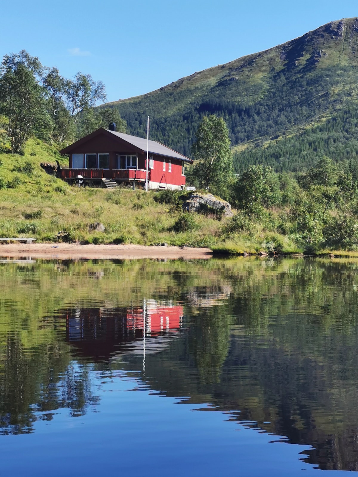 A red cabin is set against a backdrop of lush greenery and a mountain. The structure is reflected in the calm water of the lake, which features a sandy shore lined with grass. The scene captures a serene and natural environment.