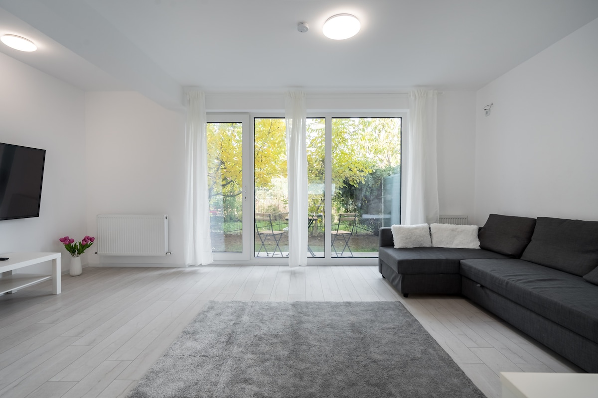 A light-filled living area features large glass doors that open to a garden view. A dark sectional sofa is positioned in the corner, with a cozy rug beneath. Potted plants add a touch of color, and a wall-mounted flat-screen television completes the space.