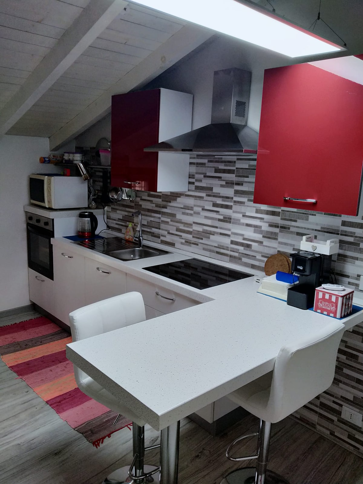 A modern kitchen features sleek cabinetry in white and red, with a contemporary countertop and bar seating. A variety of appliances, including an oven, microwave, and coffee machine, are arranged efficiently. A striped rug adds a subtle touch of color to the floor.