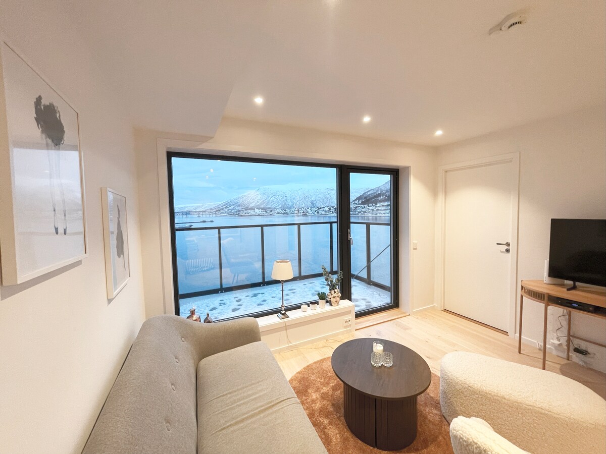 A modern living area is displayed, featuring a large window showcasing views of the fjord and surrounding mountains. A circular coffee table occupies the center, surrounded by a gray sofa and a textured armchair. Soft lighting is provided by recessed lights in the ceiling.