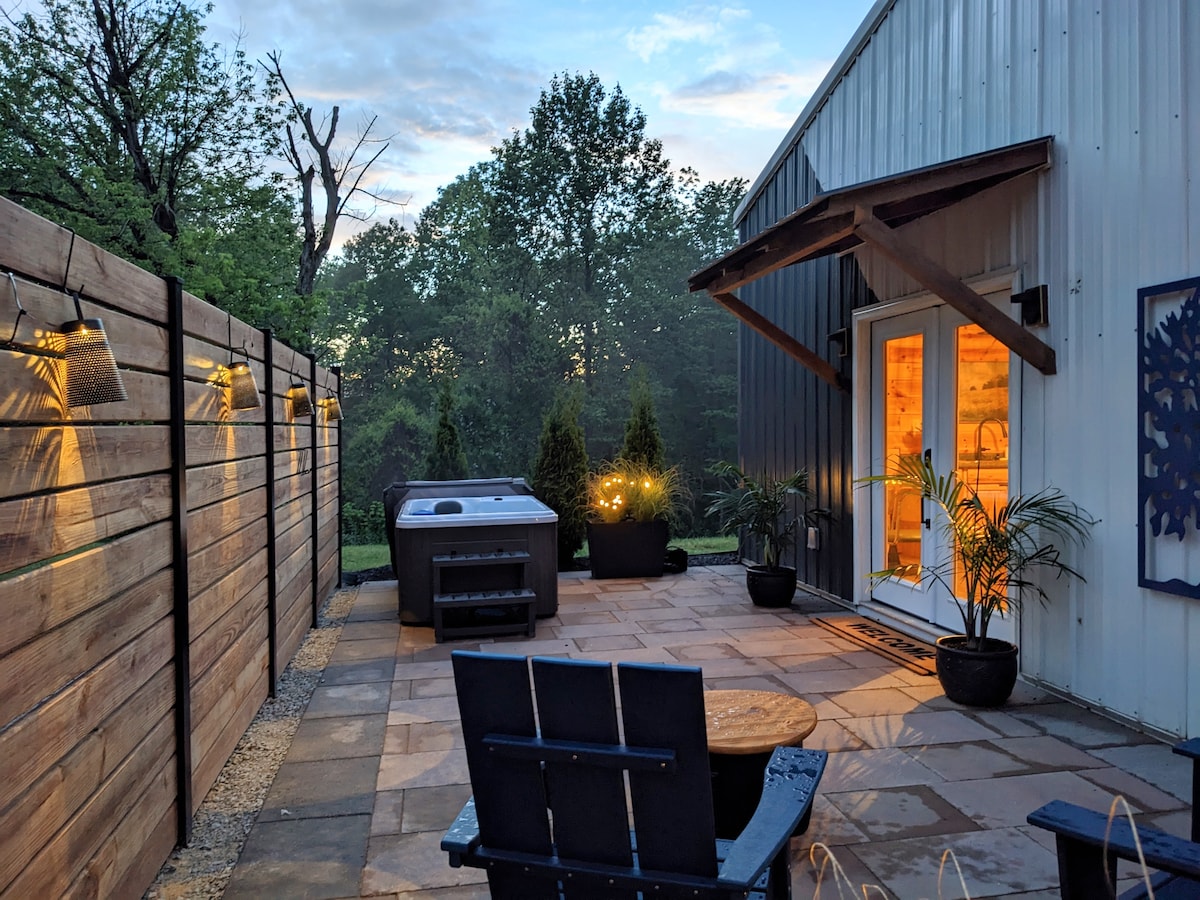 An outdoor patio area features a hot tub and a circular fire pit surrounded by black chairs. Soft lighting emanates from the privacy fence, highlighting potted plants. A backdrop of trees and a twilight sky create a serene setting for relaxation.