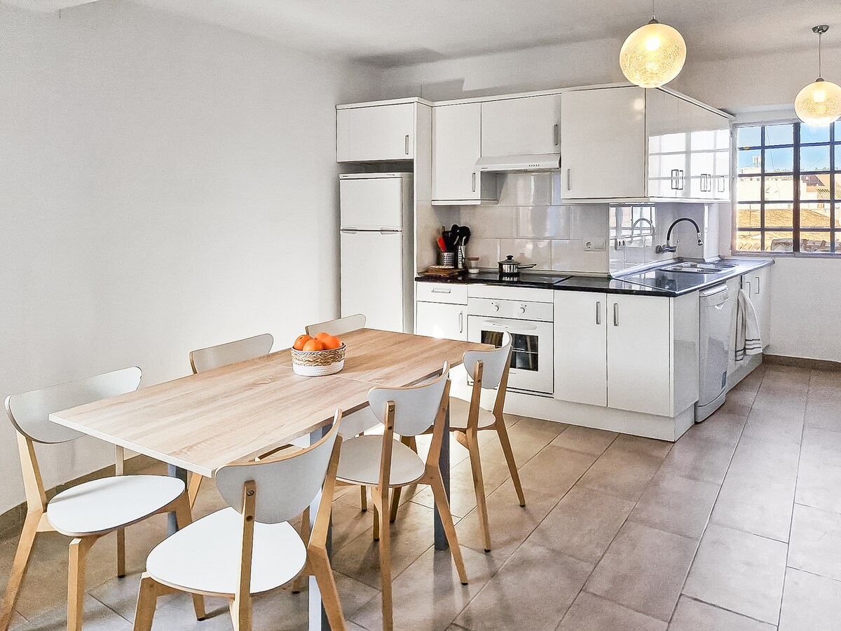 A modern kitchen is showcased with sleek white cabinetry and stainless steel appliances. A spacious wooden dining table with six light-colored chairs is positioned in the center. Natural light pours through a window, illuminating the room.