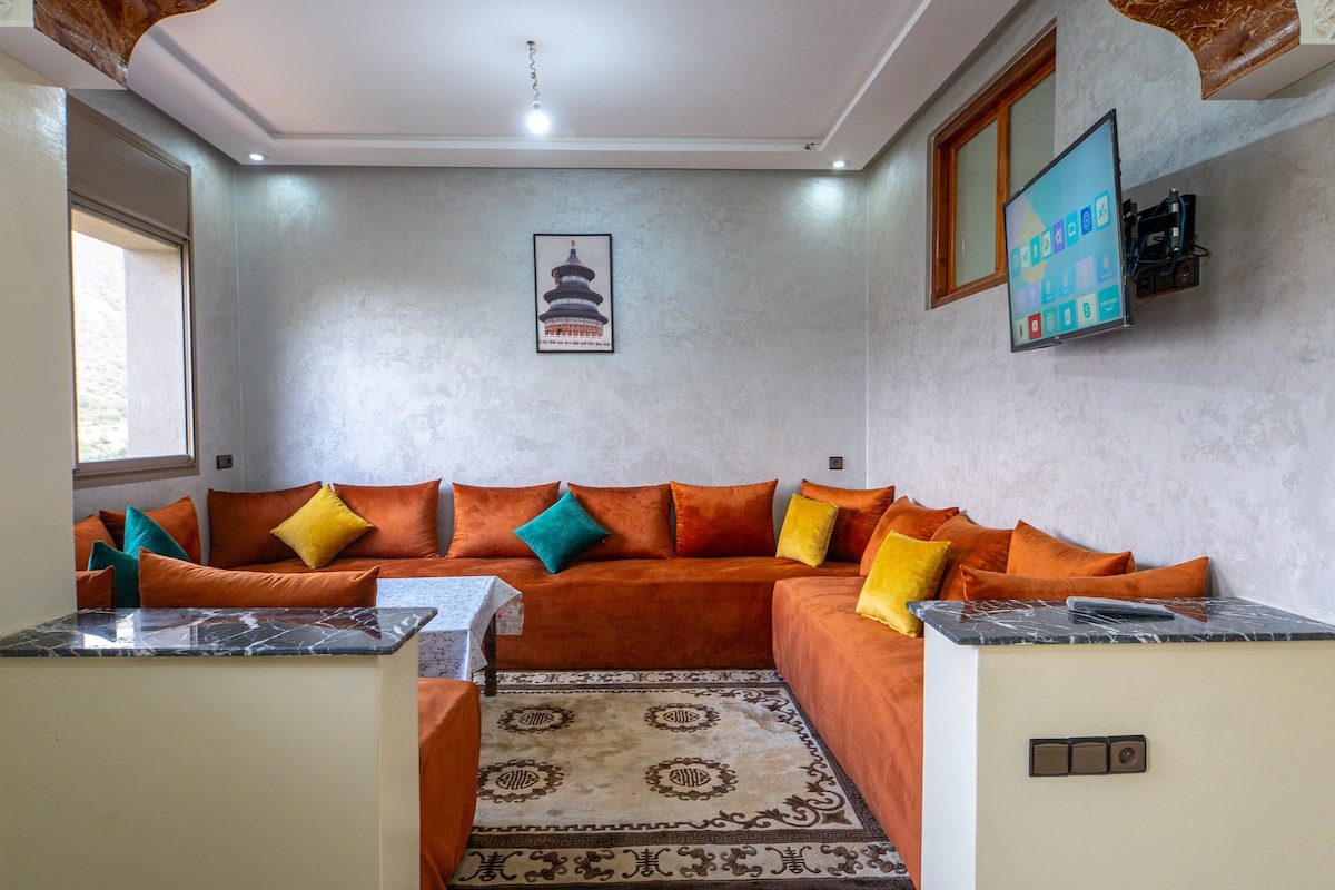A cozy lounge area features a spacious, L-shaped orange sofa adorned with colorful cushions. A low, rectangular table sits at the center, while a wall-mounted television is visible above. Natural light enters through a window, with decorative patterns on the floor adding a touch of character.