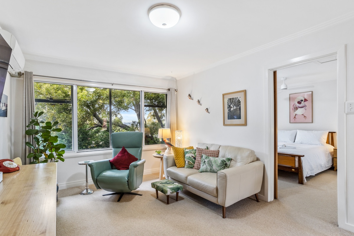 A bright living space is showcased, featuring a comfortable beige sofa and a modern green armchair. Large windows allow natural light to fill the room, framed by lush greenery outside. A small table and decorative cushions add to the inviting atmosphere.