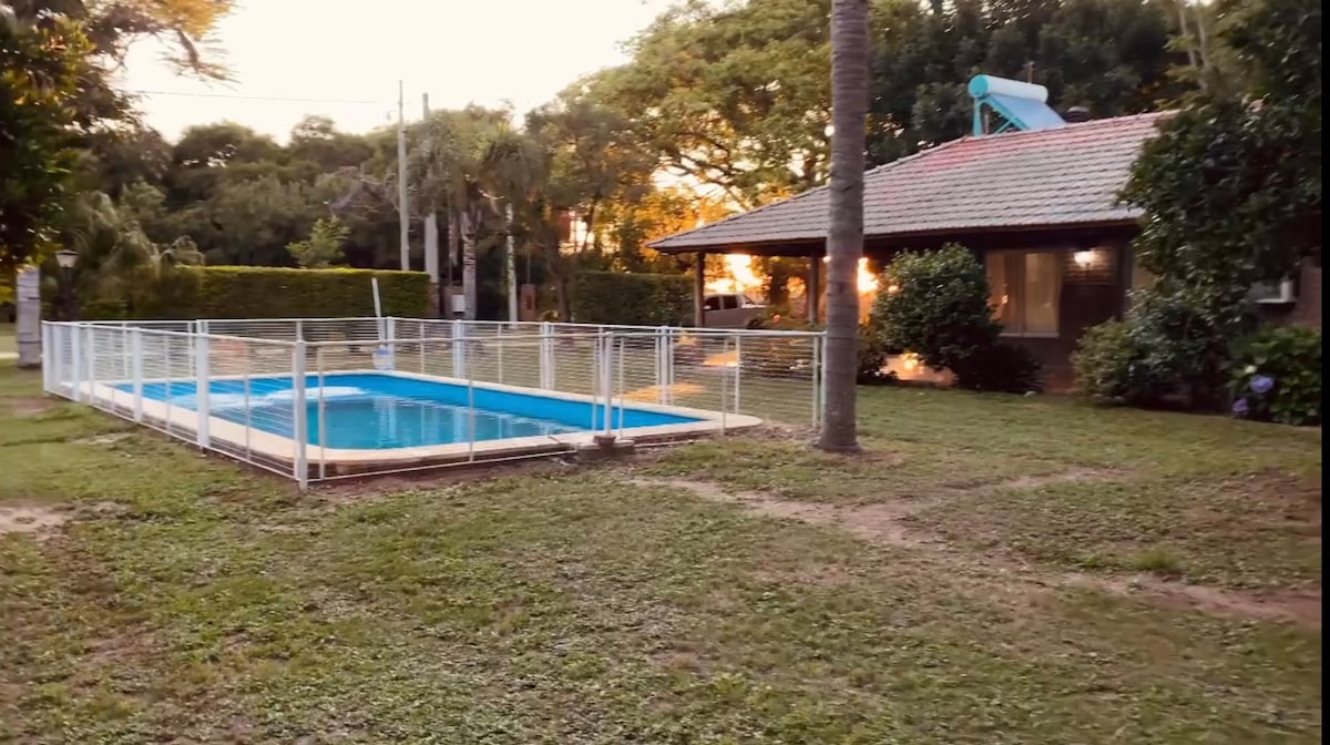 A tranquil outdoor area features a fenced swimming pool with clear blue water, surrounded by well-maintained grass. A single-story home with large windows is visible in the background, framed by trees and softly lit by the setting sun.
