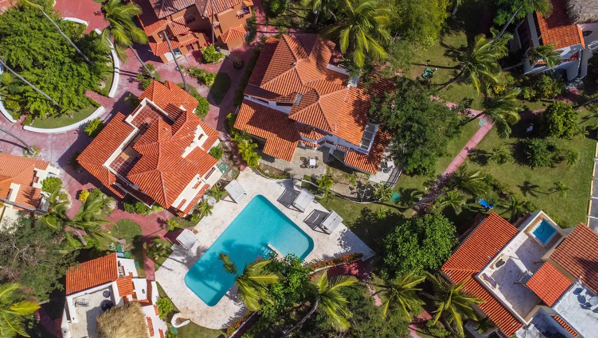 An aerial view showcases a landscaped community featuring vibrant red-roofed villas surrounded by lush greenery. In the center, a pool is nestled among palm trees, offering a refreshing area for relaxation. Pathways weave through the property, creating a welcoming environment.