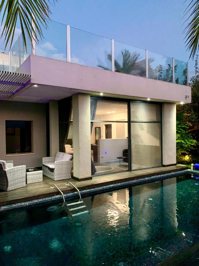 A modern villa is shown with large glass windows reflecting the evening sky. The outdoor area features a private pool with a staircase leading down, and stylish wicker seating is positioned beside it, surrounded by lush greenery.