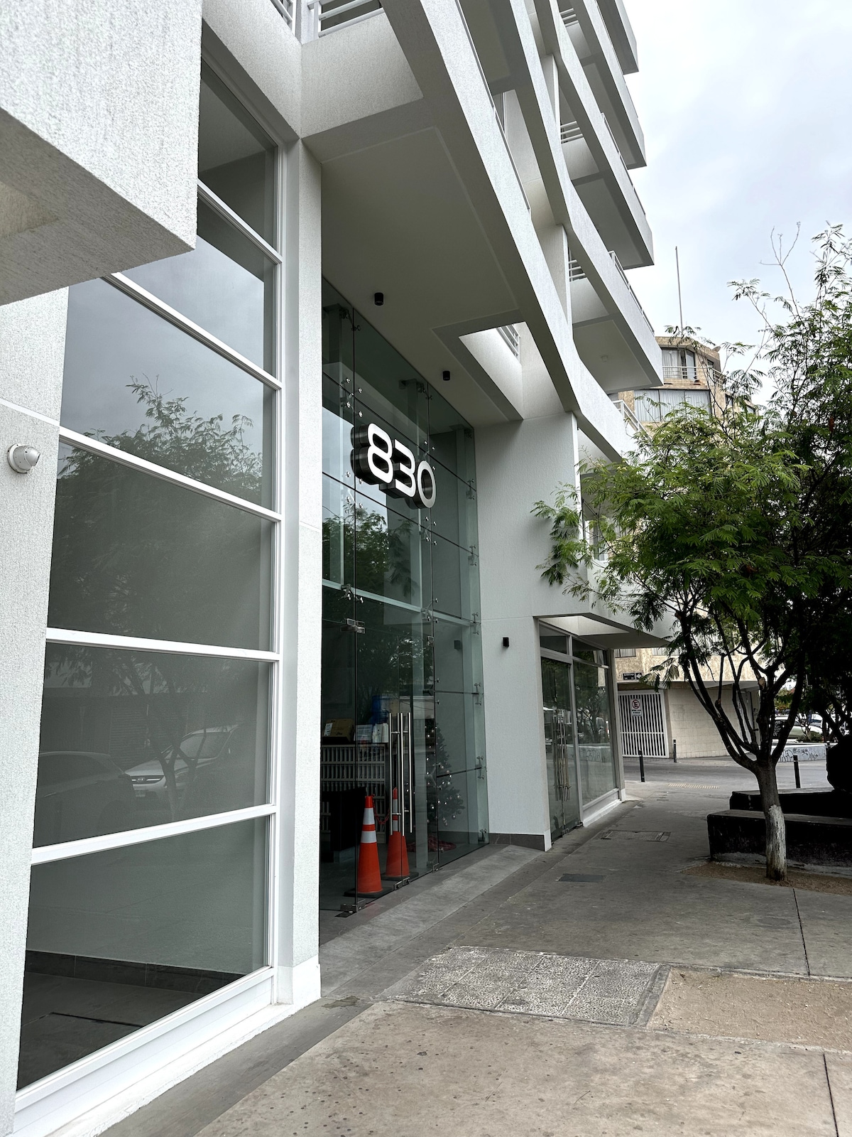 The building exterior features large glass windows that facilitate natural light. A clean white façade is complemented by a bold number display and surrounding greenery. The entrance is bordered by a pedestrian pathway and features parking cones, indicating the availability of parking facilities.
