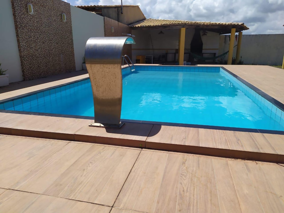A swimming pool is surrounded by wooden decking, featuring a stainless steel water slide. The pool's inviting blue water reflects the sky, with a partially shaded area visible in the background. A textured wall adds visual interest to the space.