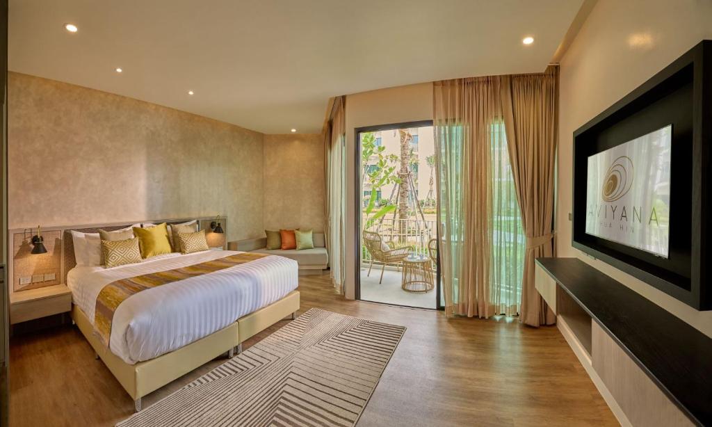 A spacious room is highlighted by a king-sized bed adorned with decorative pillows. Natural light floods in through large glass doors leading to a private outdoor seating area. Elegant curtains frame the windows, and a modern entertainment unit is mounted on the wall.