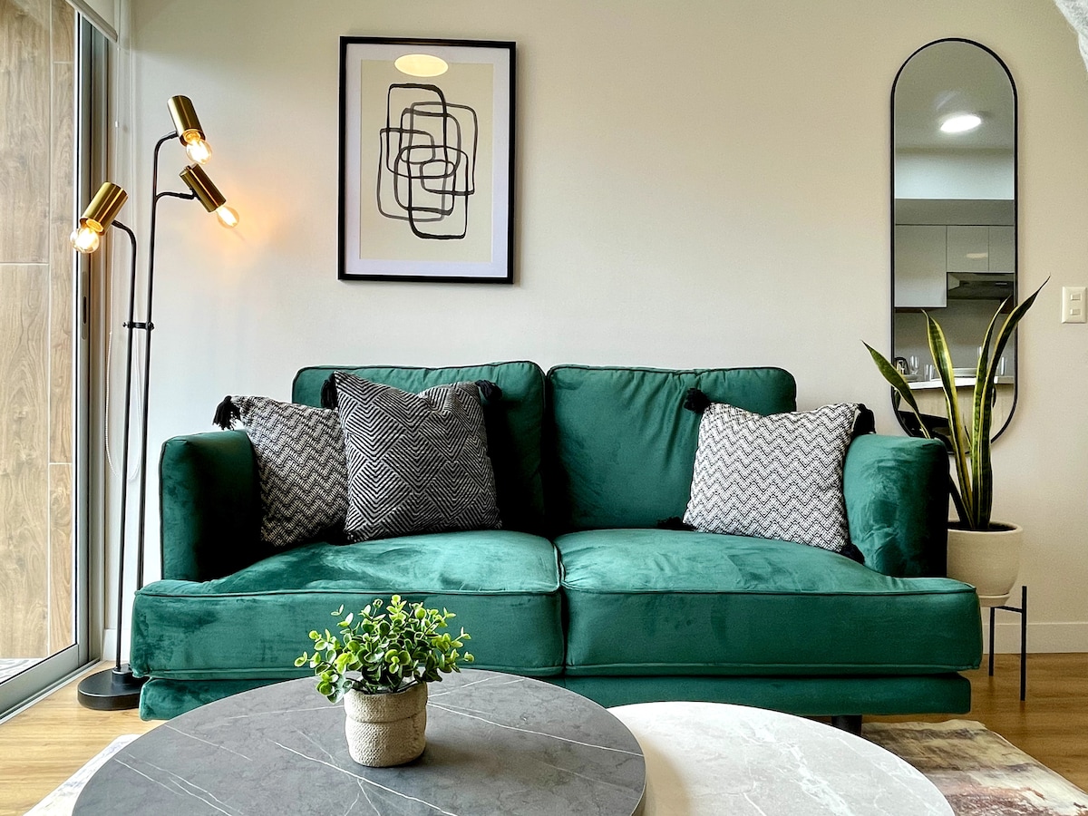 A green velvet sofa is positioned next to a round marble coffee table, which holds a small potted plant. Complementary decorative pillows add texture to the seating area, and a wall mirror reflects light. A framed artwork hangs above the sofa, enhancing the modern aesthetic.