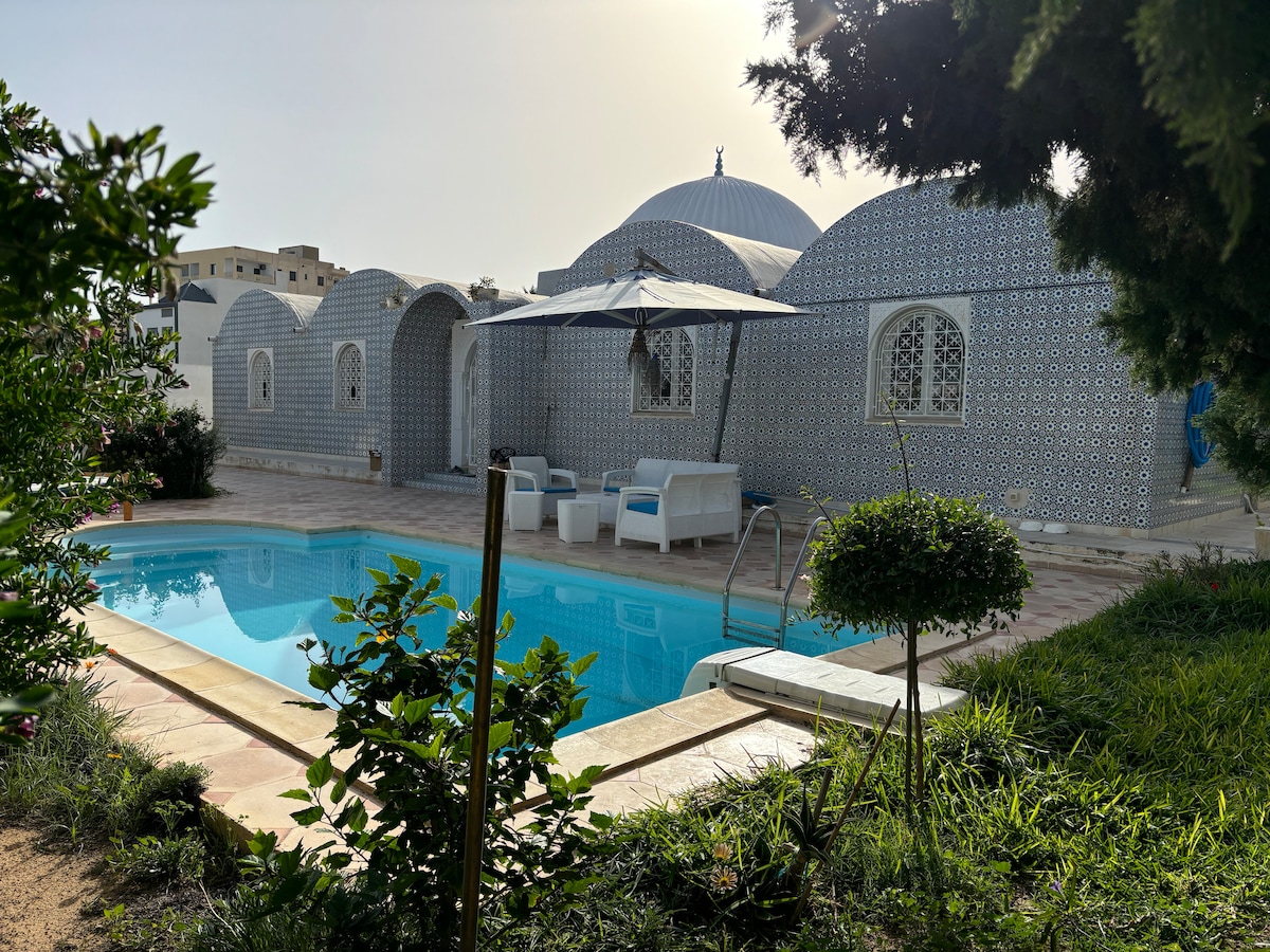 A charming property features an outdoor space with a rectangular swimming pool surrounded by greenery. Sun loungers are positioned under a large umbrella, offering a shaded area for relaxation. The unique architecture of the building is highlighted by intricate tile patterns and rounded roofs.