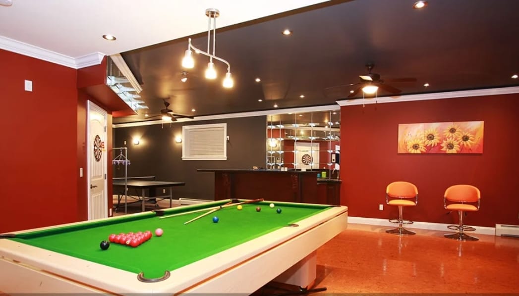 An entertainment room features a green pool table at the forefront, with colorful pool balls arranged on its surface. A wet bar with bar stools sits in the back, and additional table tennis equipment is visible in the corner. Dark red walls and artistic decor create a vibrant atmosphere.