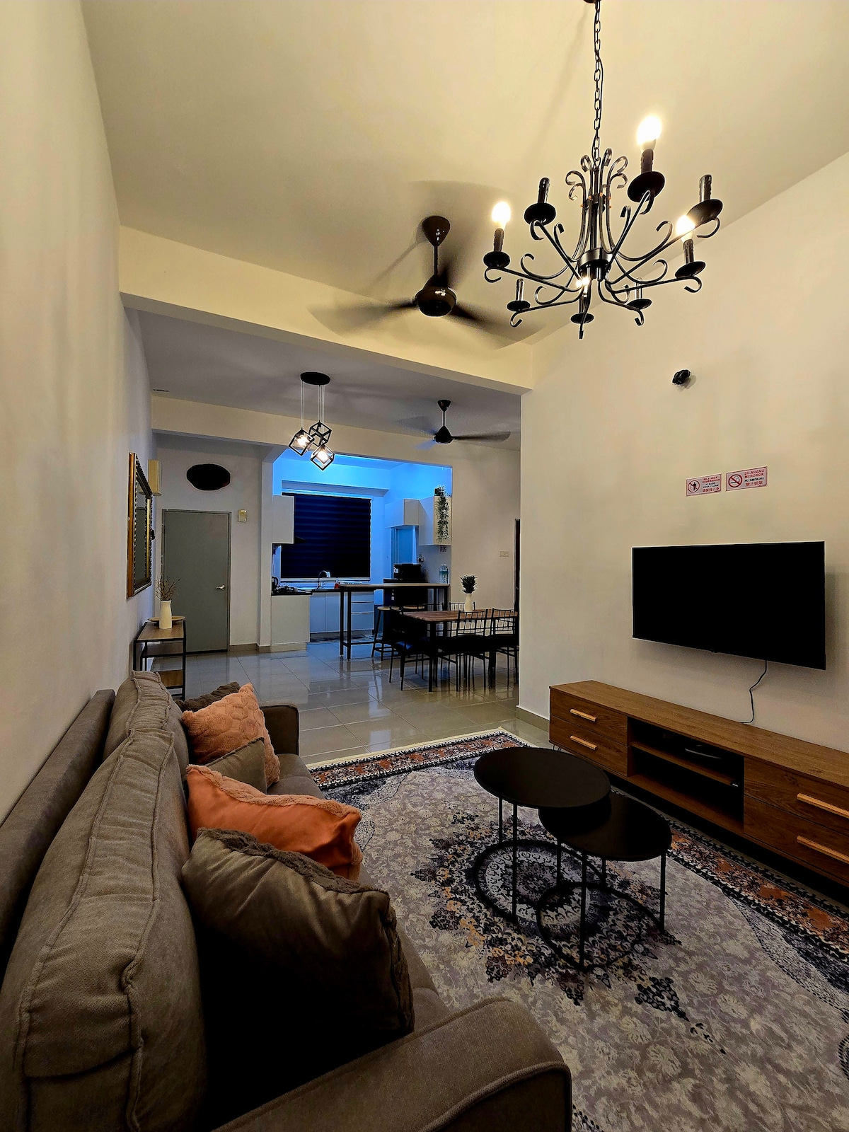 A living area is presented with a comfortable grey sofa adorned with decorative cushions. A decorative chandelier hangs from the ceiling, while a mounted television is positioned on a wooden media unit. An open view of the dining area and kitchen is available in the background.