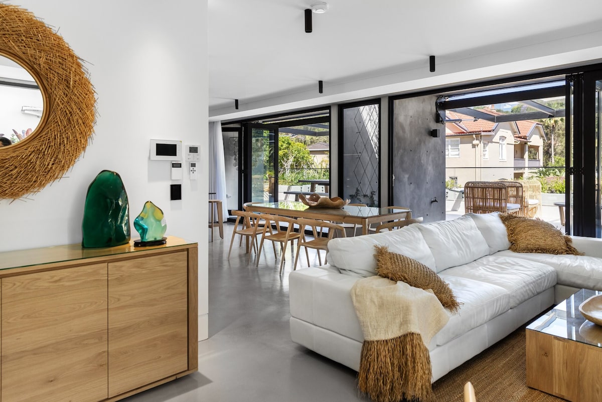 An open-plan living and dining area is captured, featuring a white sectional sofa adorned with a soft throw. A wooden sideboard and a green glass art piece add visual interest. Large glass doors invite natural light and provide access to an outdoor space.