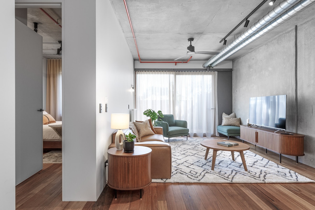 The spacious living area features large windows providing ample natural light, with a green armchair and a tan couch positioned around a wooden coffee table. A television is mounted against a concrete wall, enhancing the modern design. The warm wood flooring adds to the inviting ambiance.