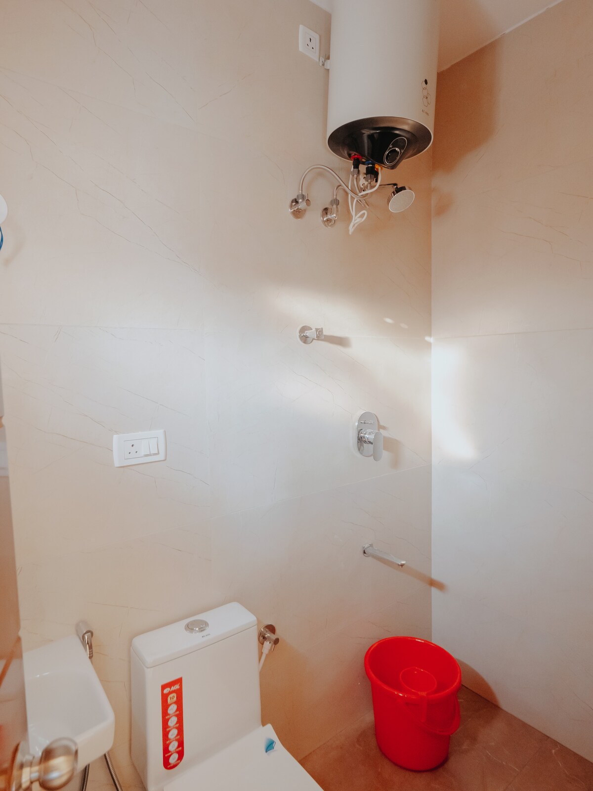 The bathroom is equipped with a modern shower area featuring dual shower heads. A toilet is positioned adjacent to the shower, and a small red bucket is placed nearby. The walls are finished with light-colored tiles, providing a clean and bright atmosphere.