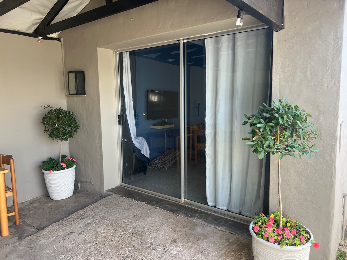 An entrance to the flat is visible, featuring a large sliding glass door. A small outdoor patio area is shown with two potted plants, each adorned with colorful flowers. A wooden table and chairs are positioned nearby, providing a welcoming space for guests.