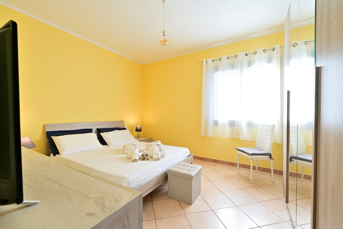 A bright yellow room is featured, showcasing a spacious bed dressed in crisp white linens. A large window allows natural light to fill the space, complemented by soft curtains. Additional seating is provided by a white chair, and a mirror reflects the room's welcoming design.