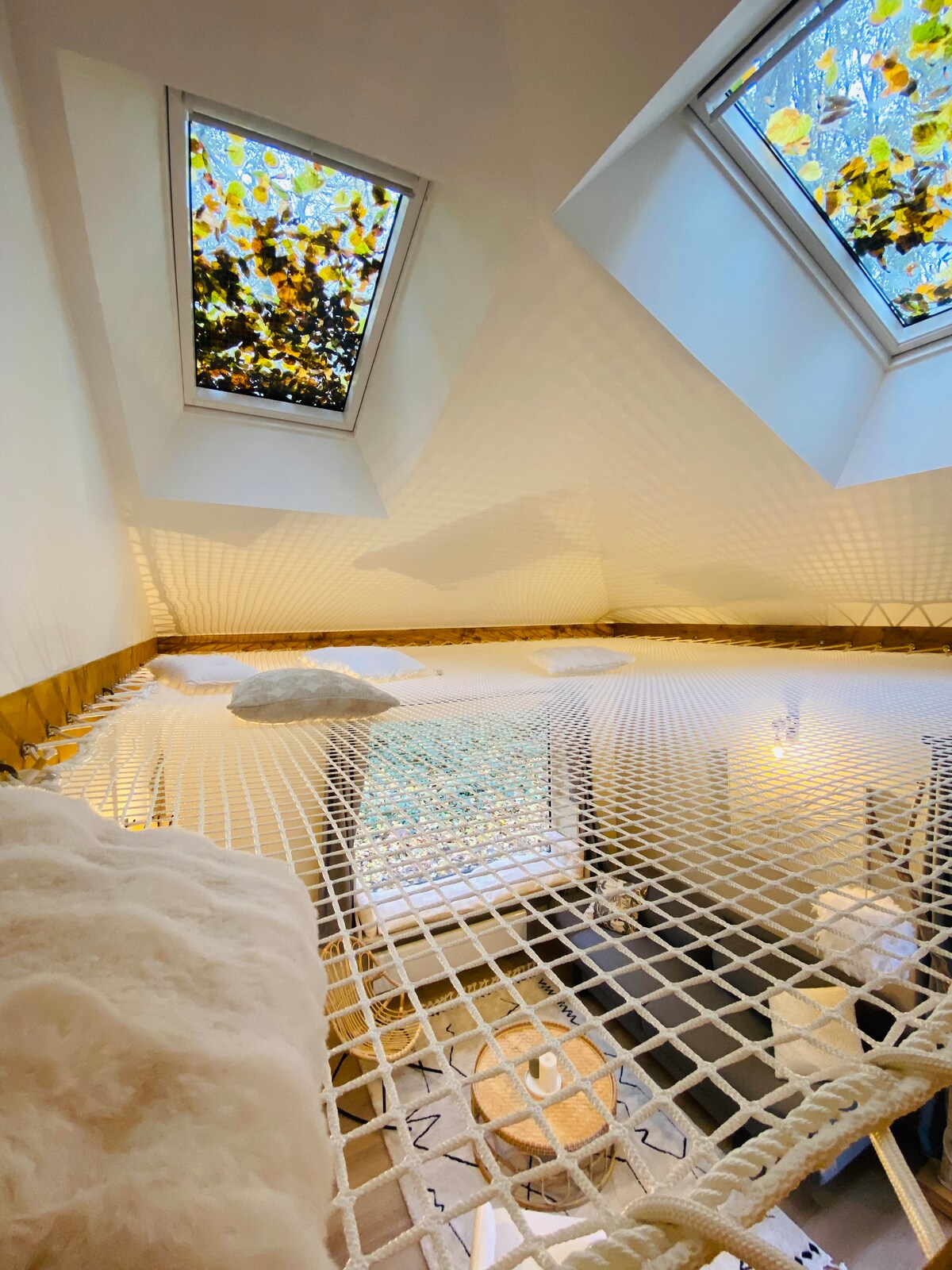 A suspended netting area is positioned beneath two angled skylights, allowing natural light and views of foliage. Soft textures in neutral tones are present, contributing to a cozy environment. The space invites relaxation and connection with nature, providing a unique resting spot.