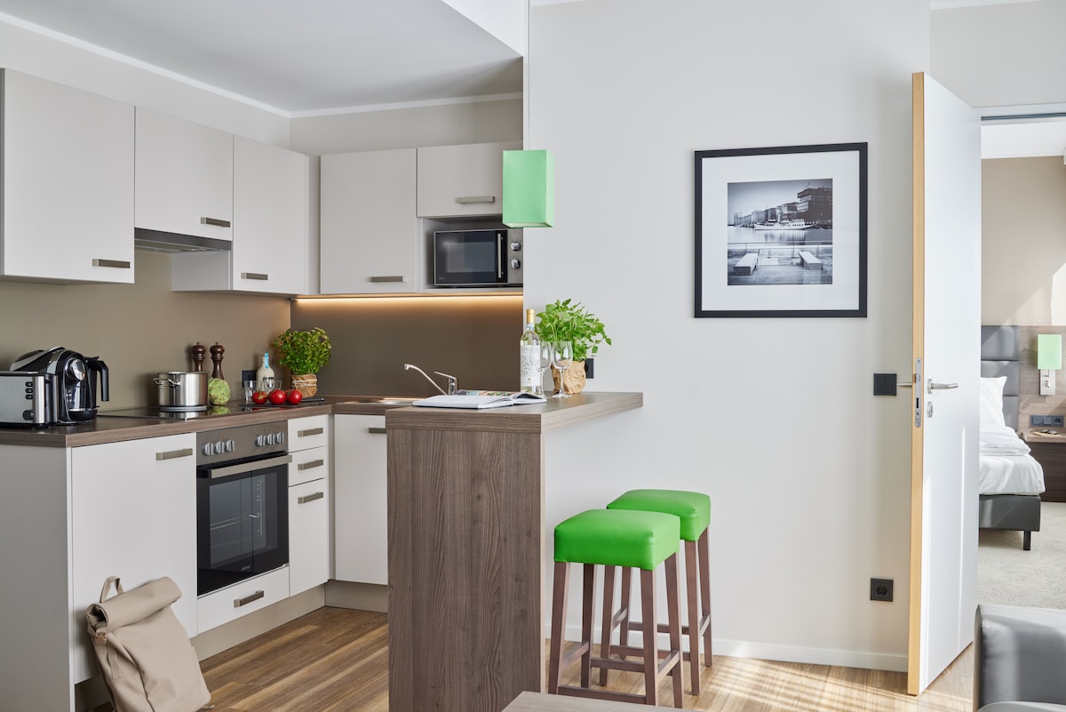 A modern kitchenette features sleek white cabinetry and dark wood accents. Essential appliances, including a stove and microwave, are arranged thoughtfully. Bar stools in a vibrant green add a pop of color to the dining area. A framed photograph is displayed on the wall.