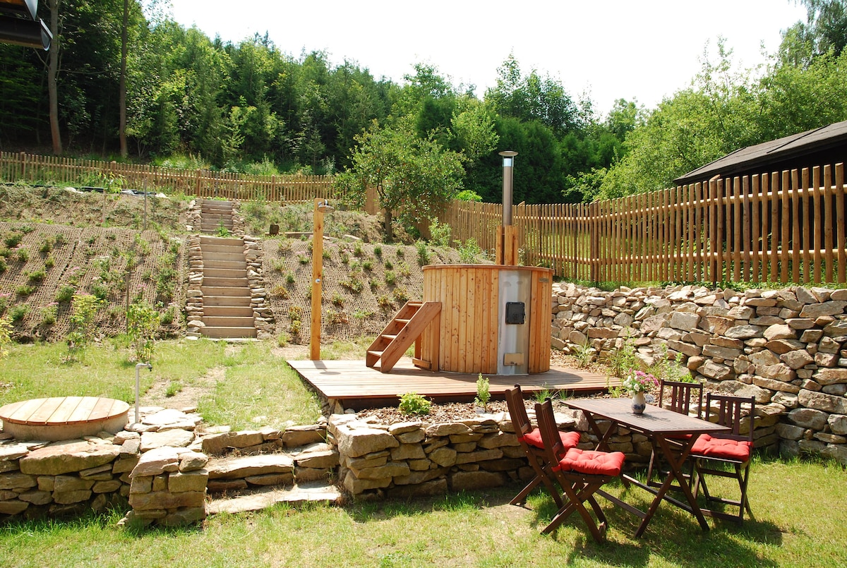 An outdoor area features a wooden hot tub situated on a spacious deck. Surrounding the tub is a wooden dining table set with four chairs, inviting relaxation. Stone steps lead to a terraced garden area, bordered by a wooden fence and greenery.