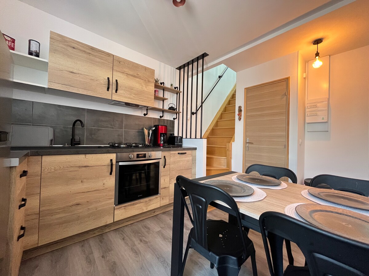The kitchen area is equipped with modern appliances and wooden cabinetry. A dining table with black chairs is positioned nearby. Natural light is seen coming in from a window. The staircase leading to the upper level is also visible.