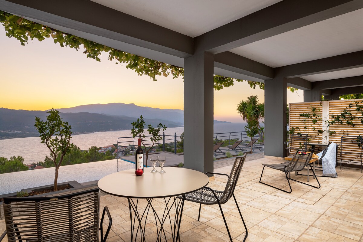 A spacious outdoor terrace is presented, featuring a round table surrounded by several chairs. The scene showcases a stunning sunset over the water, with mountains in the background. Potted plants add a touch of greenery to the setting, enhancing the overall tranquil atmosphere.