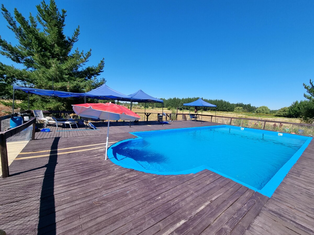 A large outdoor pool features a vibrant blue color set against a sunny backdrop. Surrounding the pool are shaded areas with tents and a few lounge chairs. The wooden deck provides ample space for relaxation, with open views of the natural landscape in the distance.