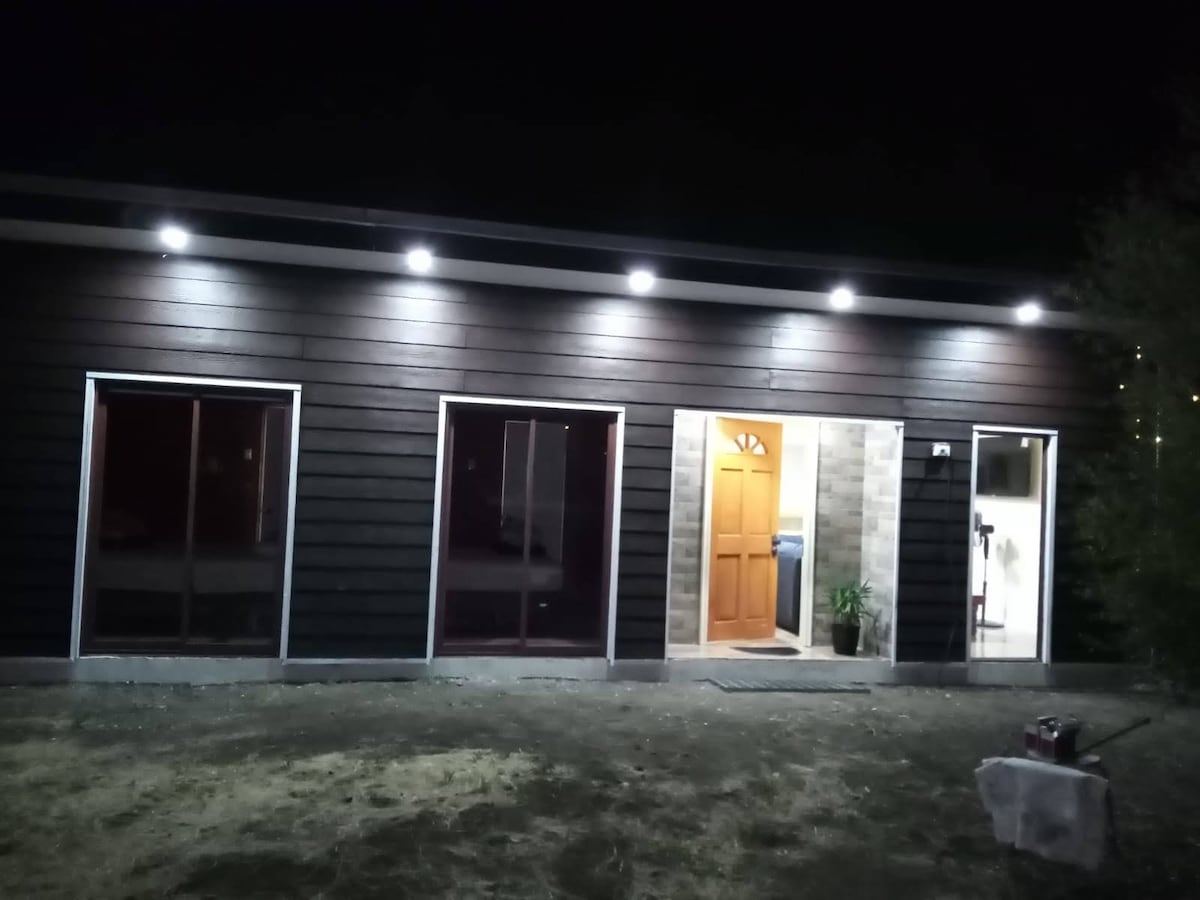 The exterior of the cabin is illuminated by soft lighting, showcasing a simple yet modern design. Large windows provide glimpses of the interior, with a welcoming front door leading inside. The surrounding area features natural landscaping, creating a serene atmosphere.