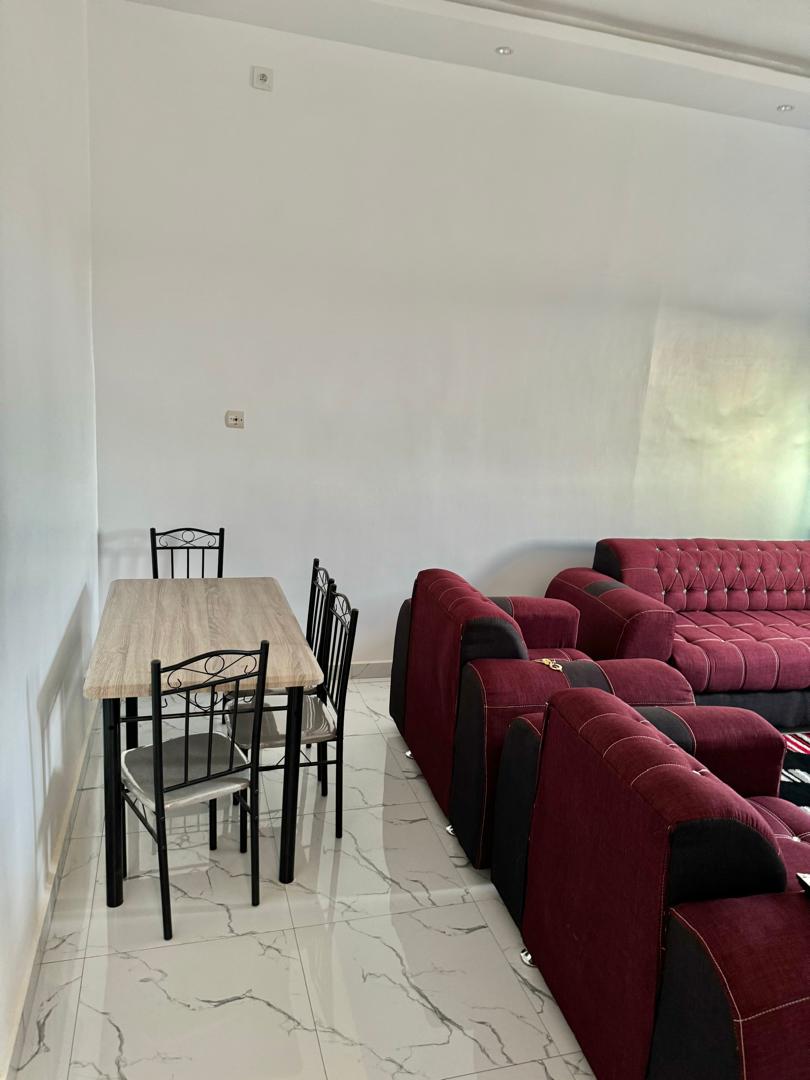 A dining table with four black metal chairs is placed on a polished tiled floor. A spacious red sofa with tufted upholstery is situated nearby, creating a comfortable seating area. Soft natural light illuminates the room from a window, enhancing the airy ambiance.