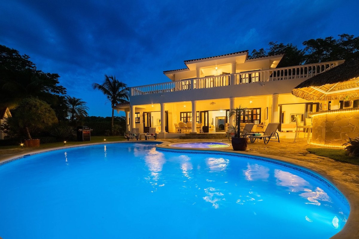 The villa is illuminated at night, highlighting its two-story structure and a spacious outdoor area. A bright blue pool glistens in the foreground, accompanied by a separate jacuzzi. Surrounding palm trees and the tropical garden create a serene atmosphere.